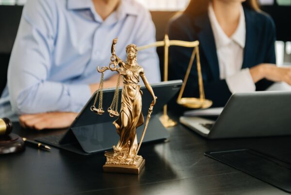 Top Myths About Pre-Settlement Funding – Debunked. Image of a gold statue of Justice sitting on a desk in front of two lawyers working on laptops and tablets.